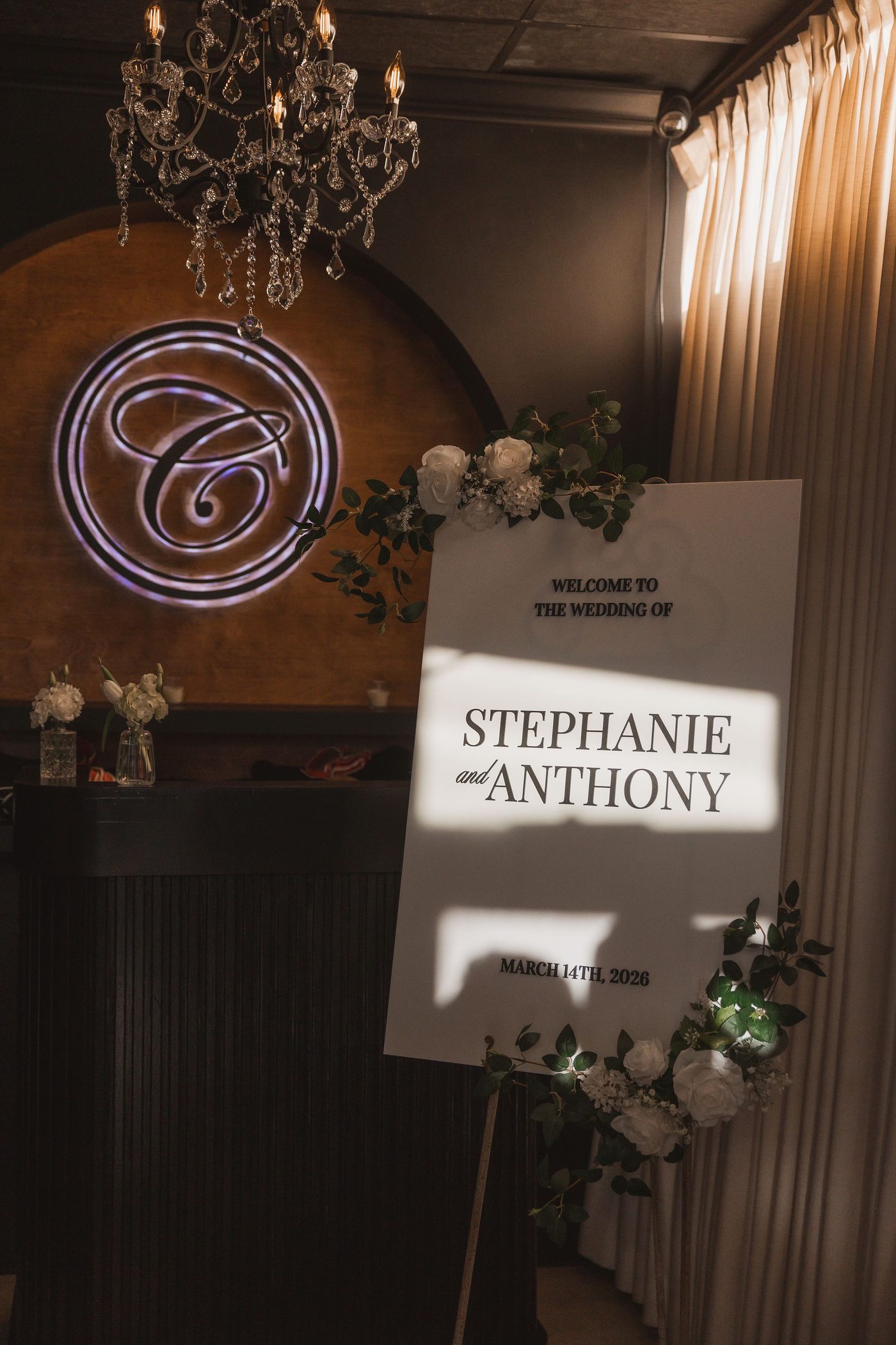 A wedding welcome sign framed by white roses and eucalyptus beneath the chandelier in the Giavanna Room at Clarizio's.